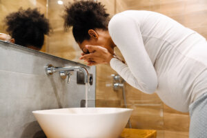 A pregnant woman is washing her face over a sink in a bathroom, practicing self care.