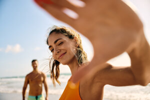 A couple is on the beach with waves in the background. The woman is holding her hand up to the camera lens and smiling while the man is several feet behind her.